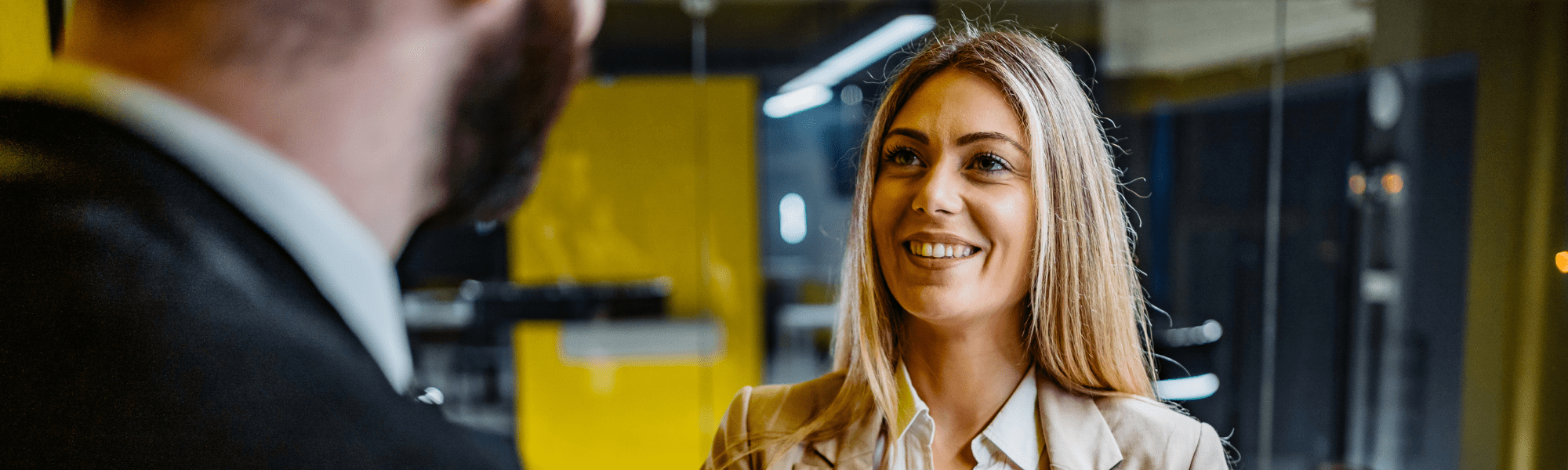 Close-up of a woman with blonde hair wearing a light brown jacket meeting a businessman out of focus in the foreground