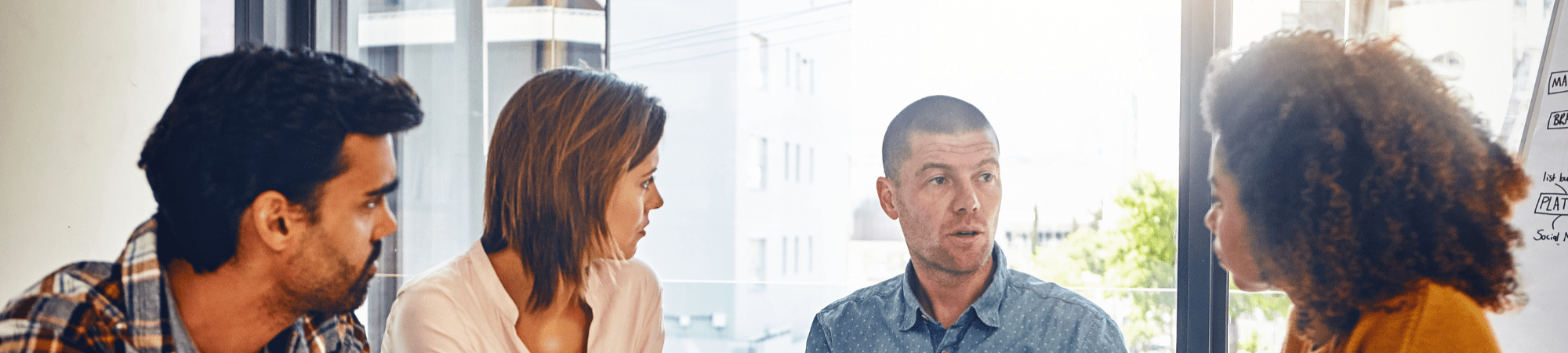 Man seated at a table discussing things with his team, two women and a man, all dressed in smart casual clothes