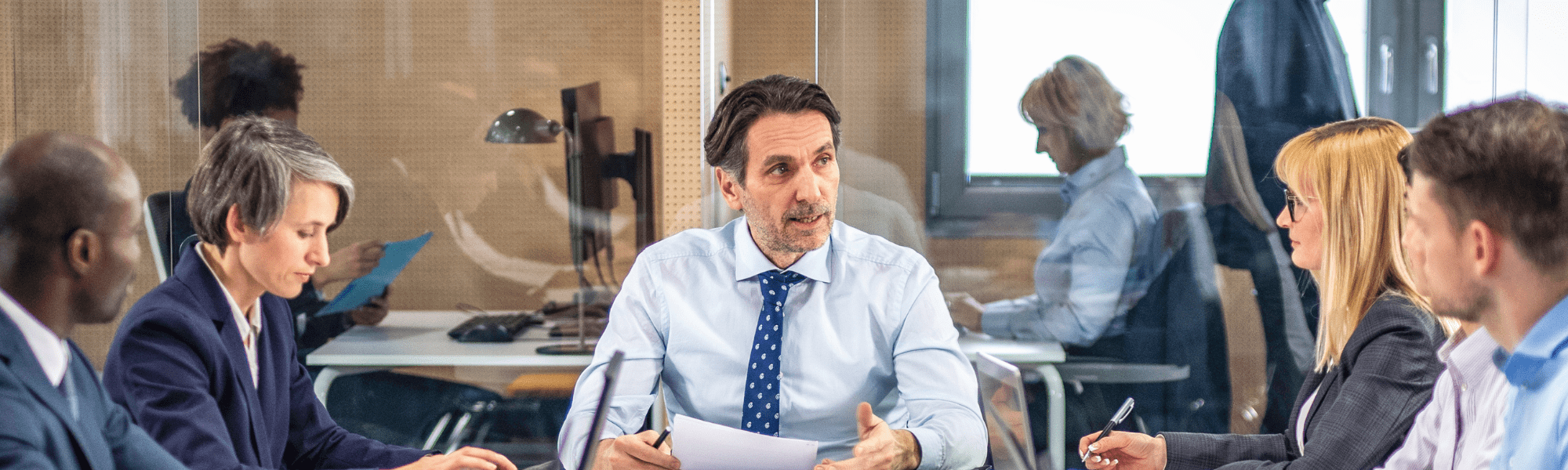 Male executive wearing a shirt and ties seated at the head of a table meeting his team