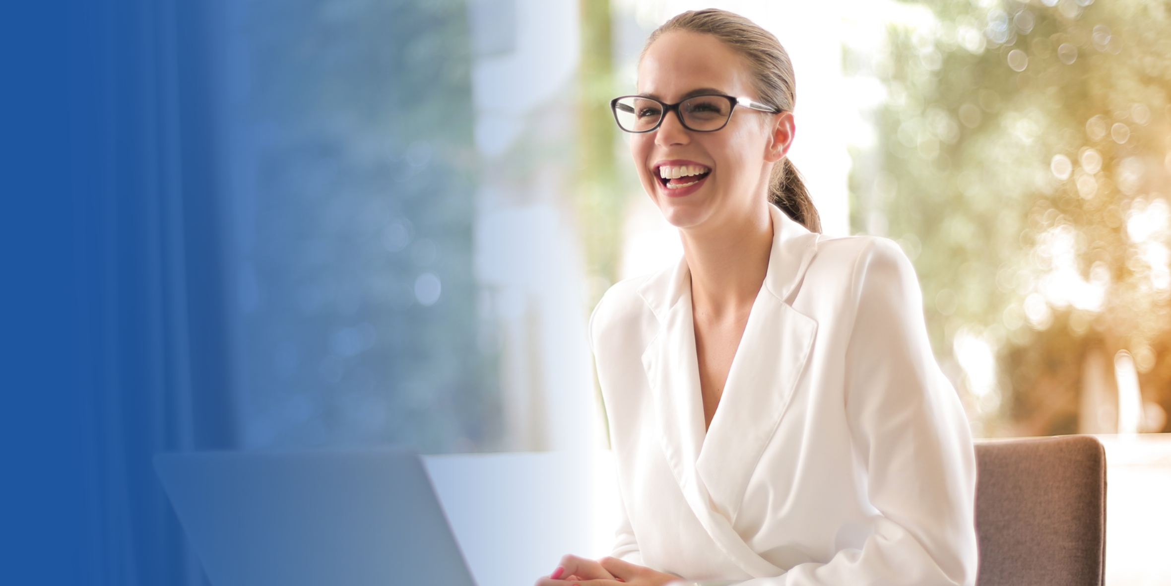 Professional businesswoman wearing white blouse sitting at a desk, with an open laptop and smiling