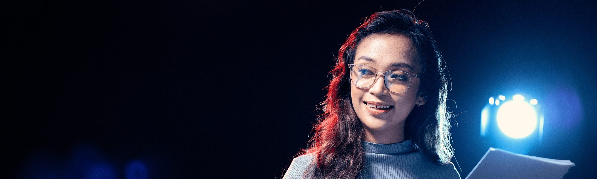 Woman backlit by a spotlight holding a script while presenting on stage 